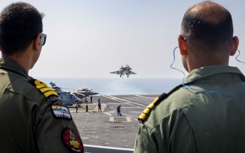 Two service members of the Indian Navy watch a Rafale fighter arrested landing on the Charles de Gaulle aircraft carrier