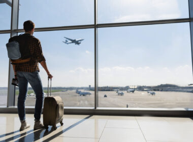 Young man is standing near window at the airport and watching plane before departure. He is standing and carrying luggage. Focus on his back