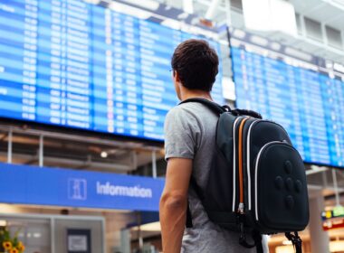 Young,Man,With,Backpack,In,Airport,Near,Flight,Timetable