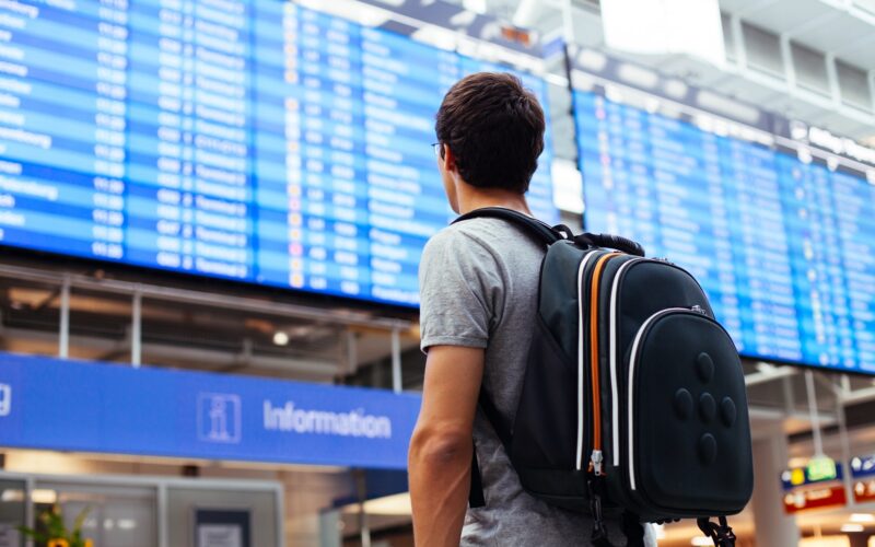 Young,Man,With,Backpack,In,Airport,Near,Flight,Timetable